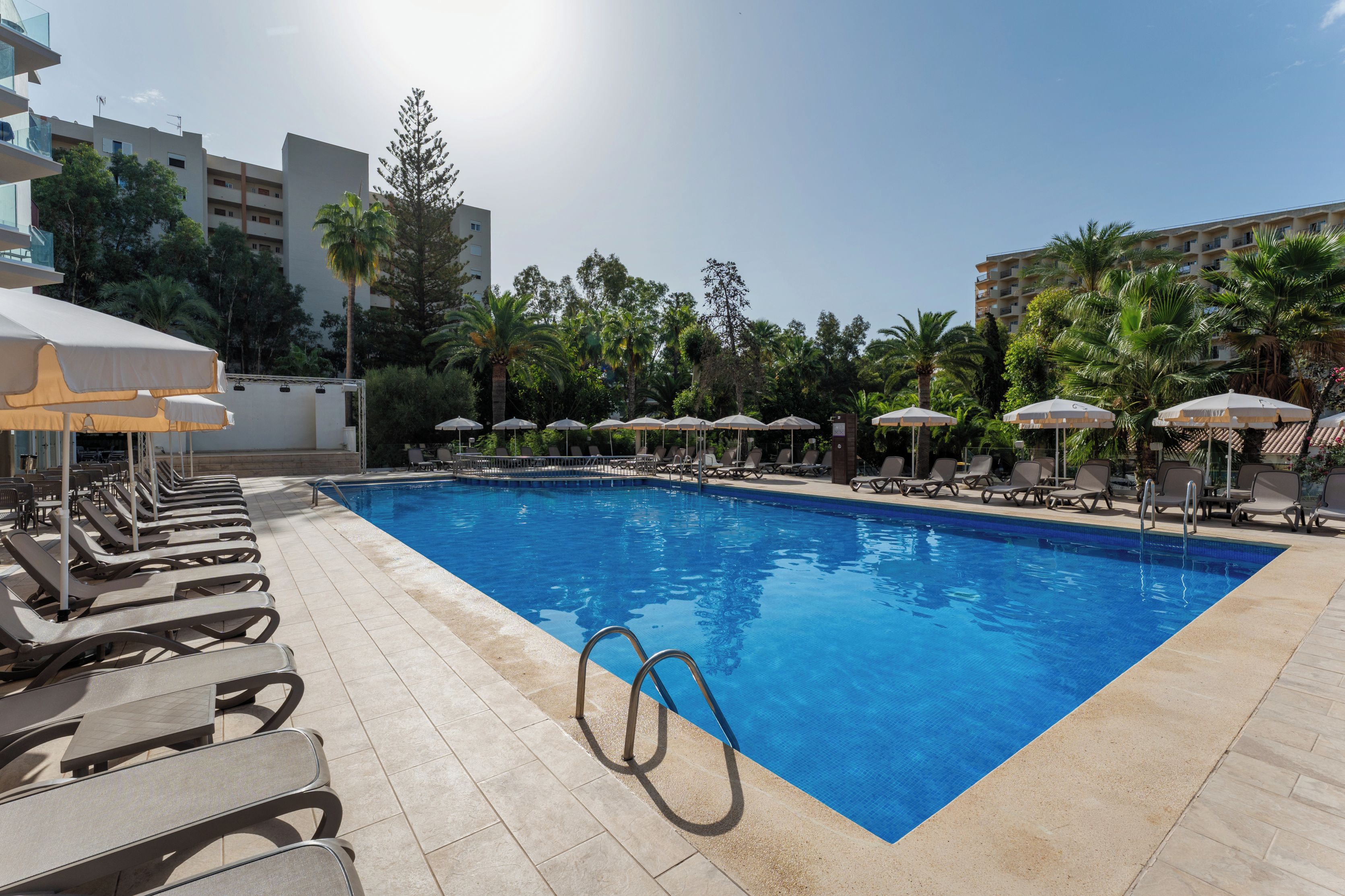 A swimming pool with sun loungers and umbrellas, surrounded by palm trees and buildings.