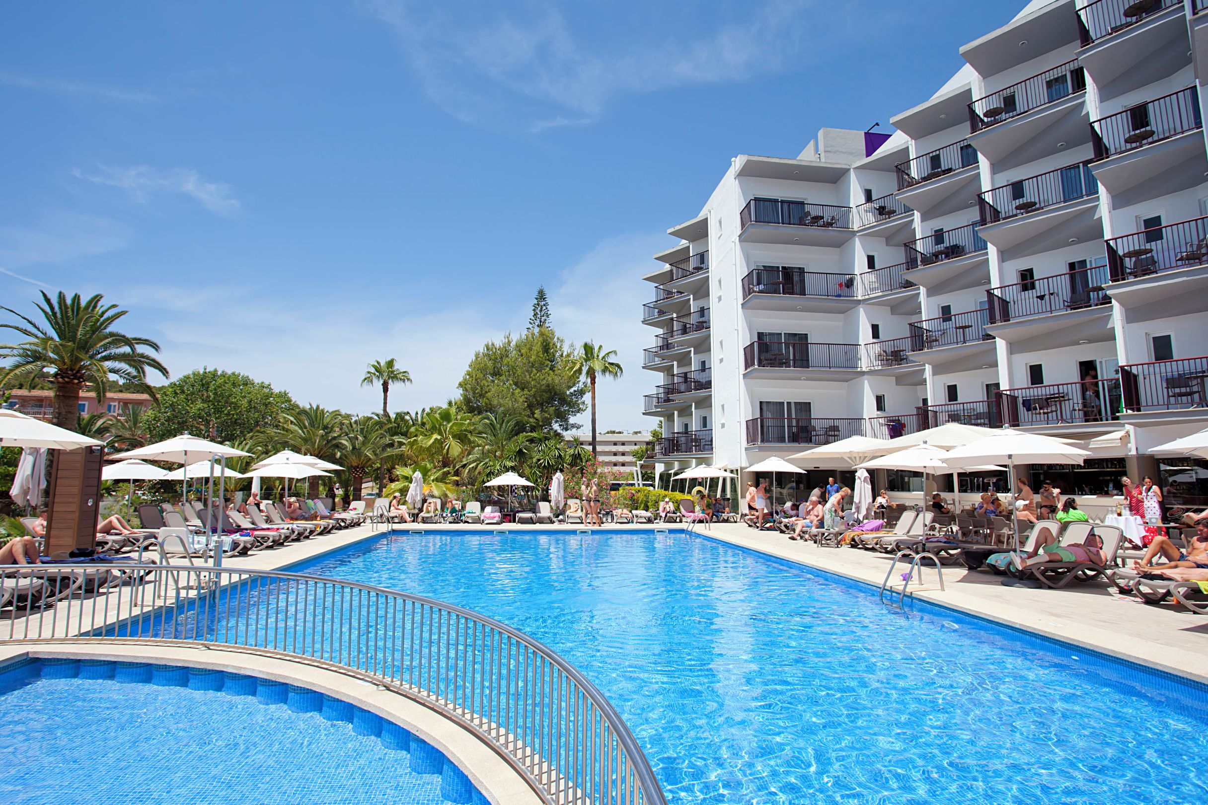 Une piscine entourée de transats et de parasols, elle-même bordée d’un immeuble à plusieurs étages doté de balcons.