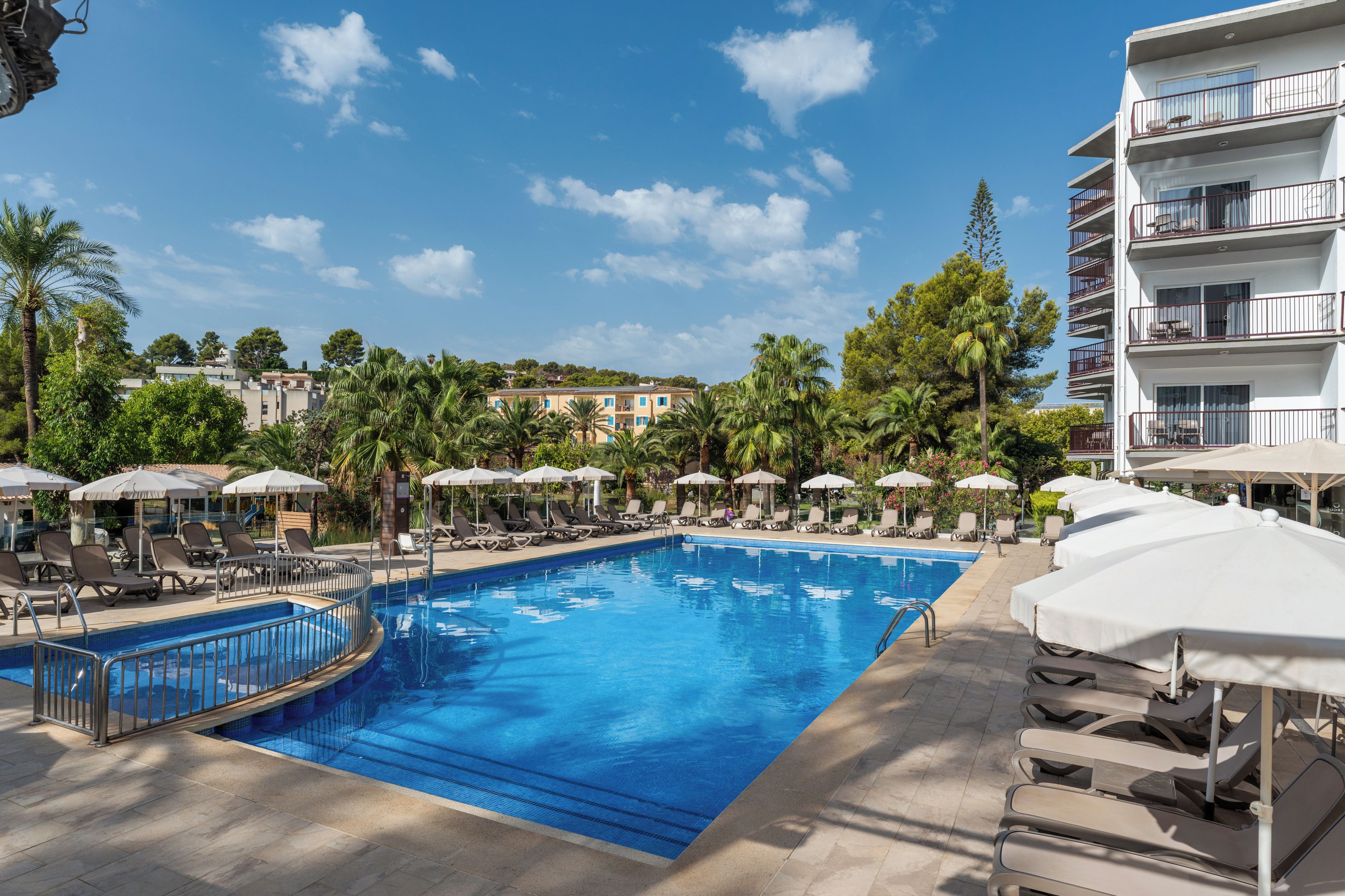 Una piscina con tumbonas y sombrillas, rodeada de palmeras y un edificio de varios pisos.