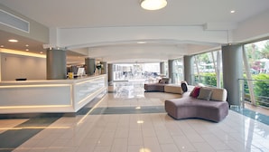 A spacious lobby with a reception desk, a curved seating area with sofas, and large windows offering views of the outdoors. - FERGUS Bermudas (Calvia)