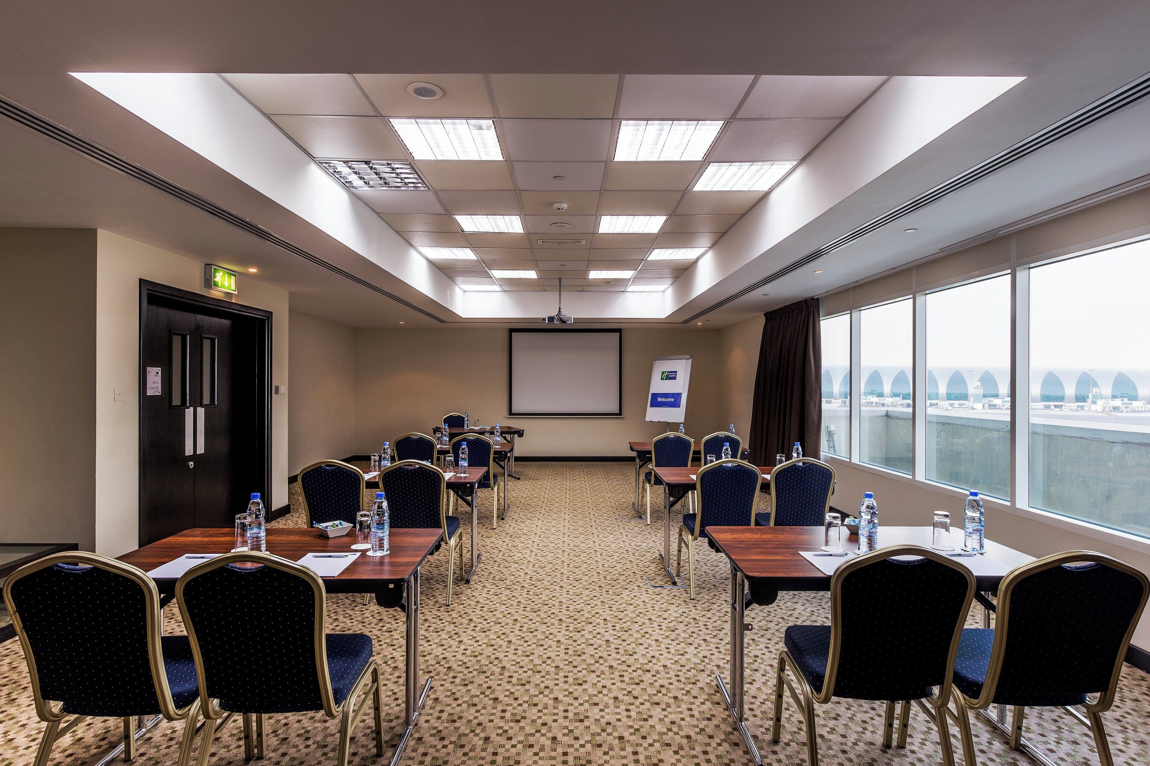 A conference room with a projector screen, a 'Welcome' sign, and a view of the airport through large windows.