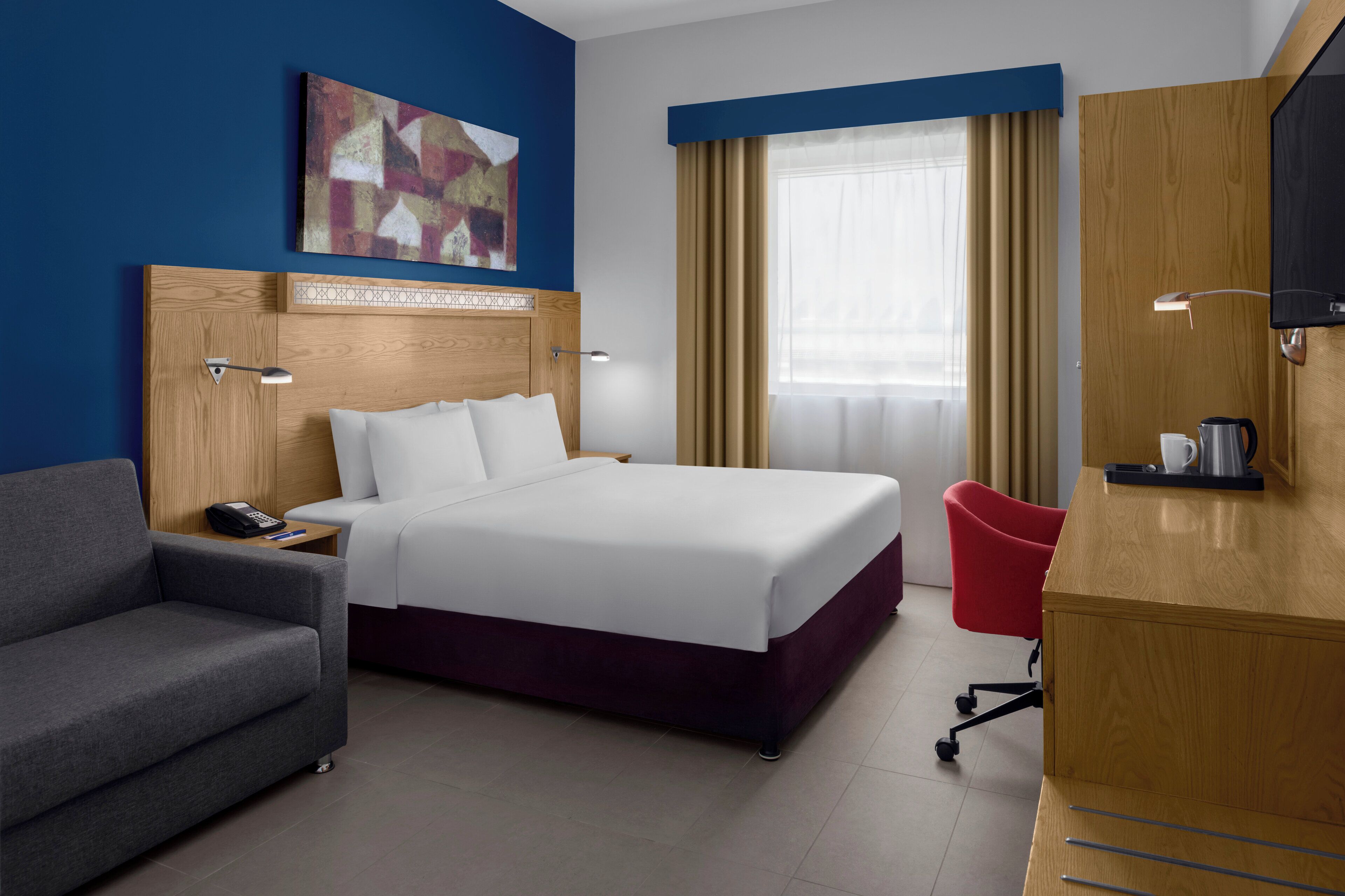 A hotel room with a large bed, a desk, a red chair, a sofa, a painting on the wall, and a window with curtains.