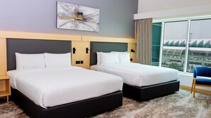 Quarto de hotel com duas camas, uma janela ampla com vista para a cidade, um quadro abstrato moderno e uma cadeira azul.