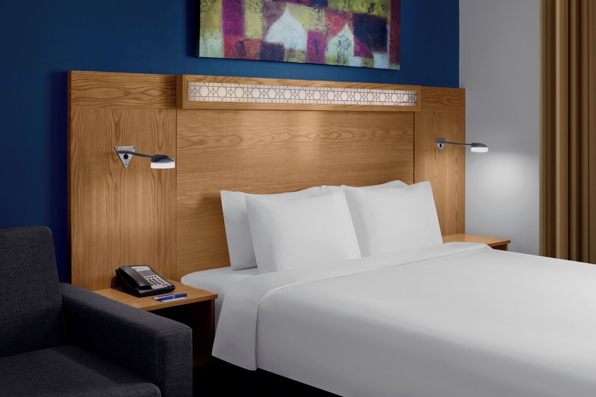 Une chambre d’hôtel avec une tête de lit en bois, du linge de lit blanc, un bureau avec un téléphone et une lampe.