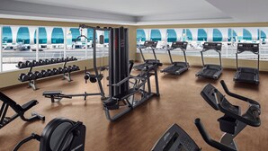 Uma academia com esteiras, halteres e aparelhos de musculação, com amplas janelas que oferecem vista para um aeroporto.