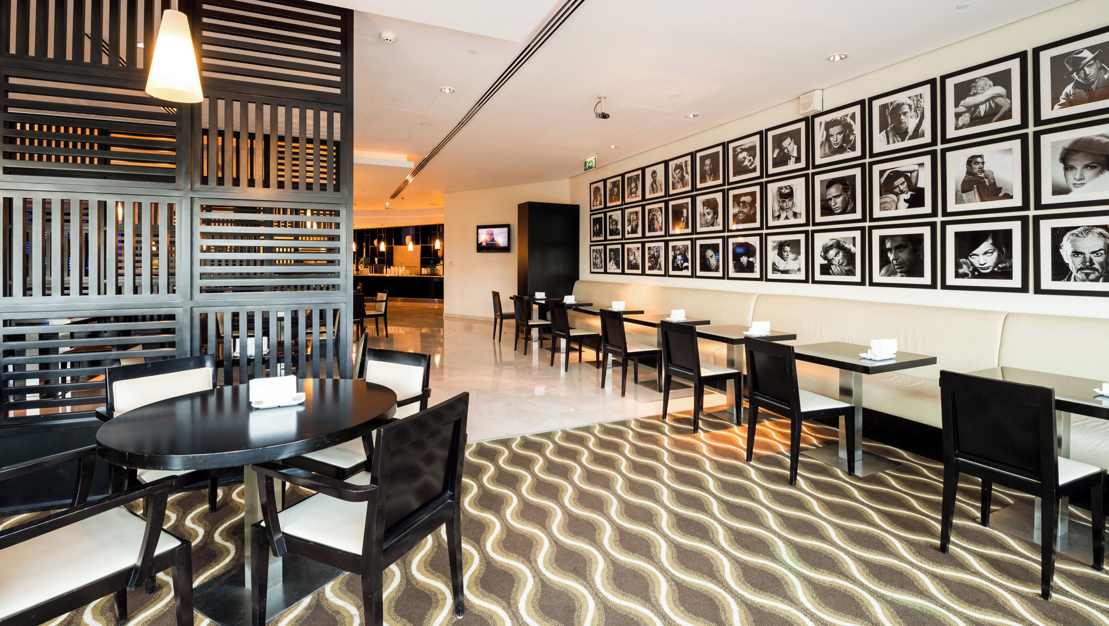 A hotel lobby with a modern design, featuring a patterned carpet, a variety of seating options including tables and chairs, and a wall adorned with framed photographs.