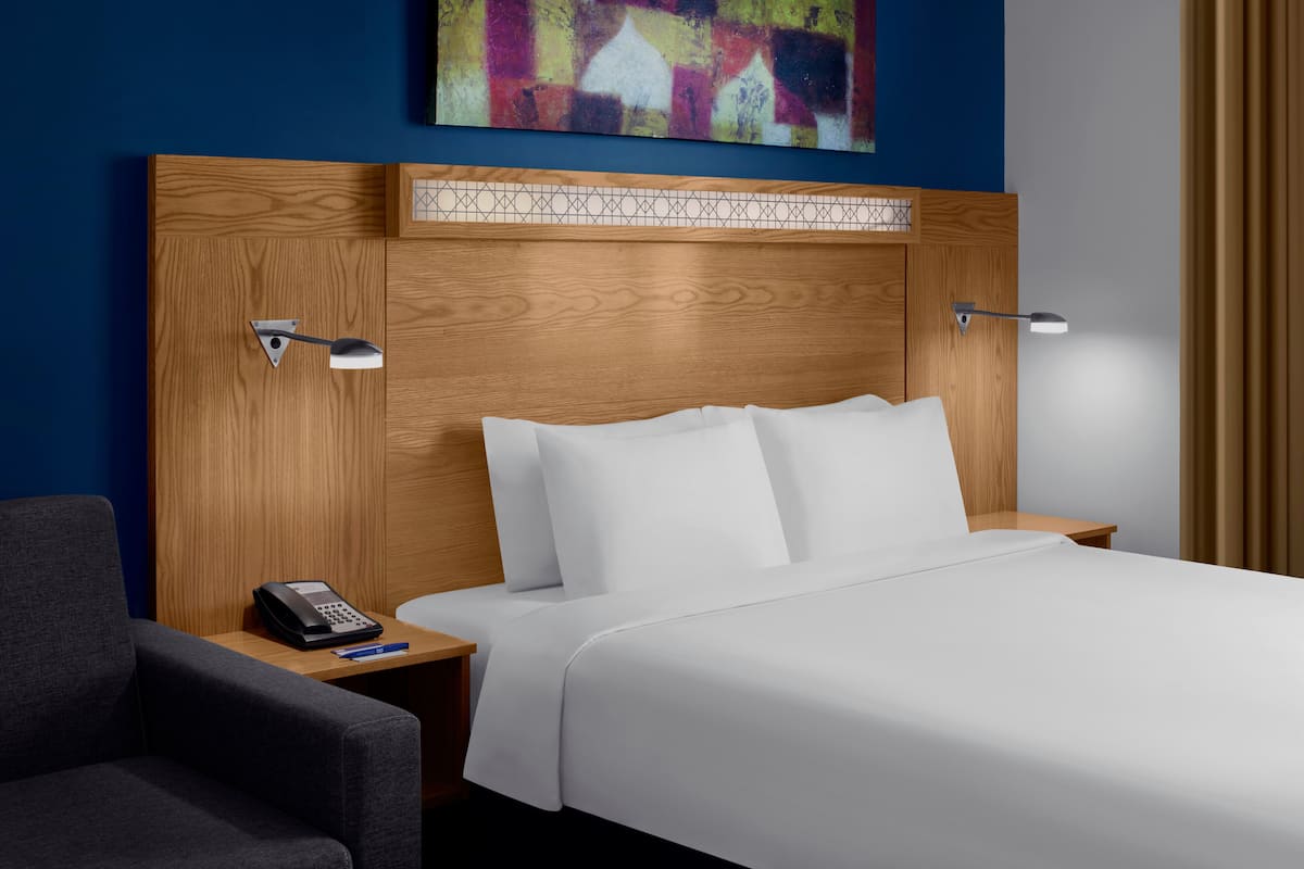 Une chambre d’hôtel avec une tête de lit en bois, du linge de lit blanc, un bureau avec un téléphone et une lampe.