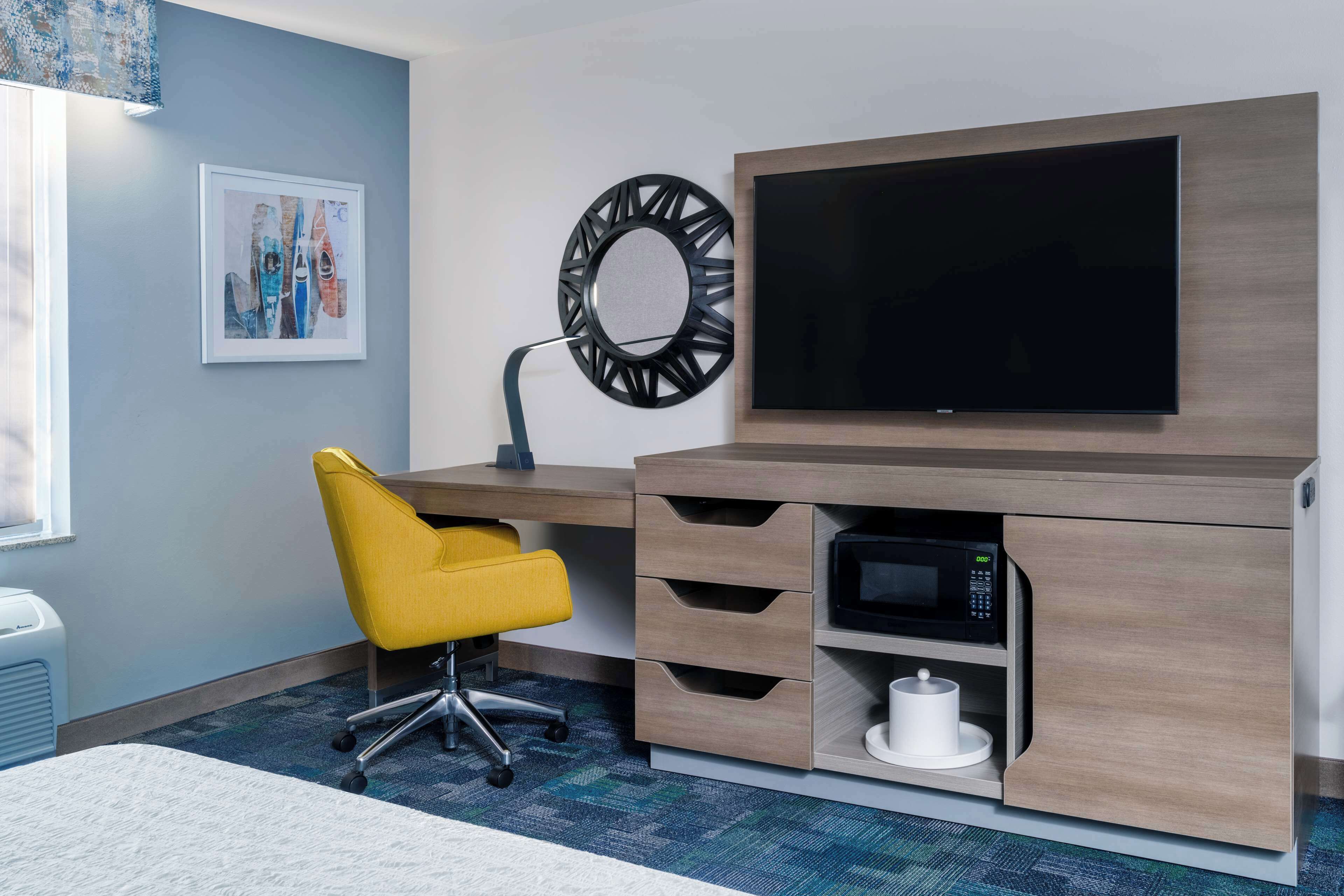 Une chambre d’hôtel dotée d’un bureau moderne, d’un téléviseur à écran plat, d’un four à micro-ondes et d’une chaise jaune.