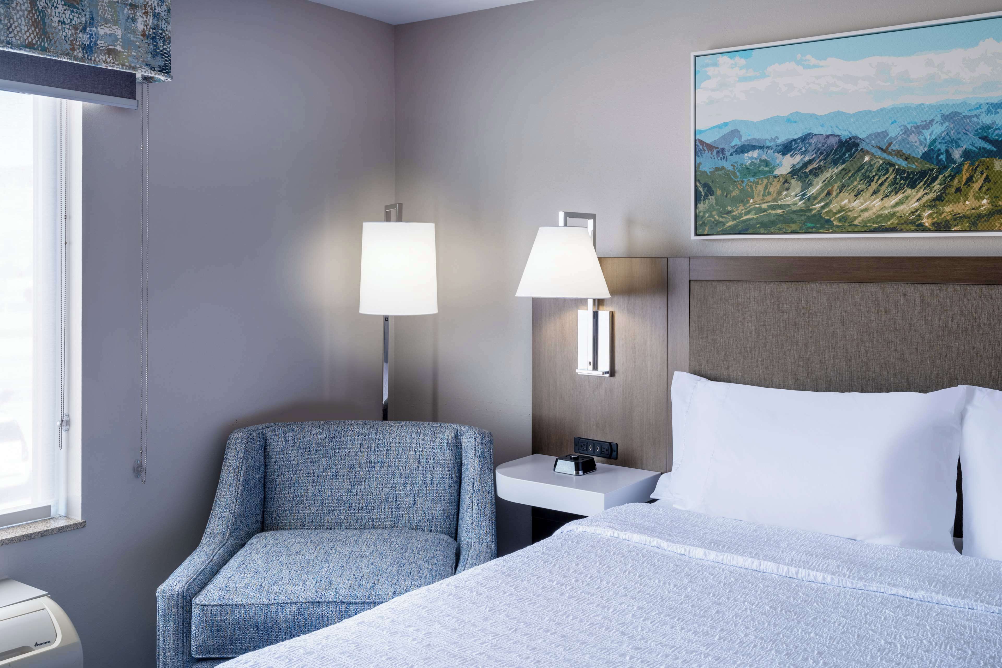 A hotel room with a bed, a chair, a nightstand, a lamp, and a painting of mountains.