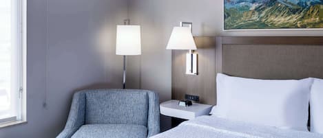 Ett hotellrum med en säng, en stol, ett nattduksbord, en lampa och en tavla som föreställer berg.