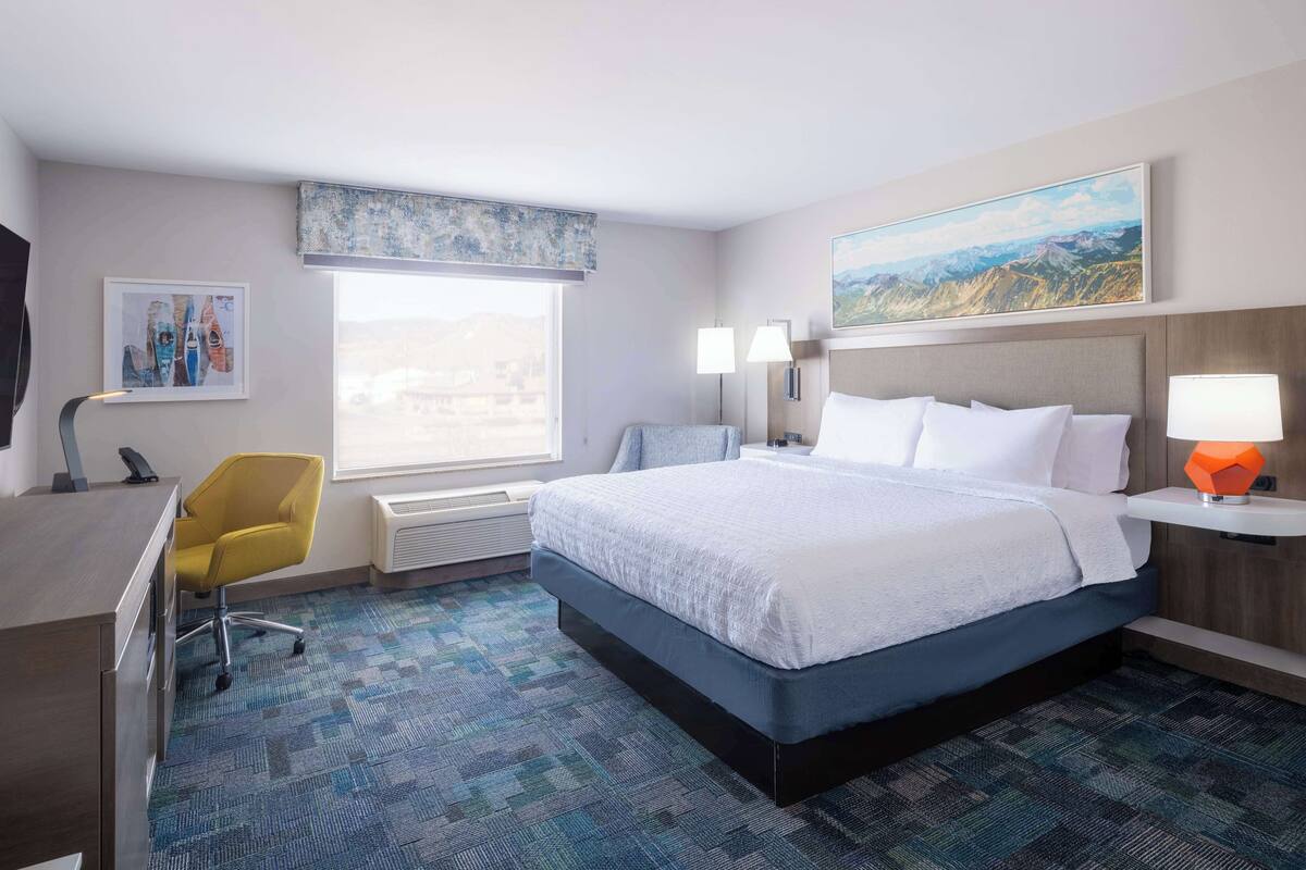 A hotel room with a bed, desk, chair, and a large window.