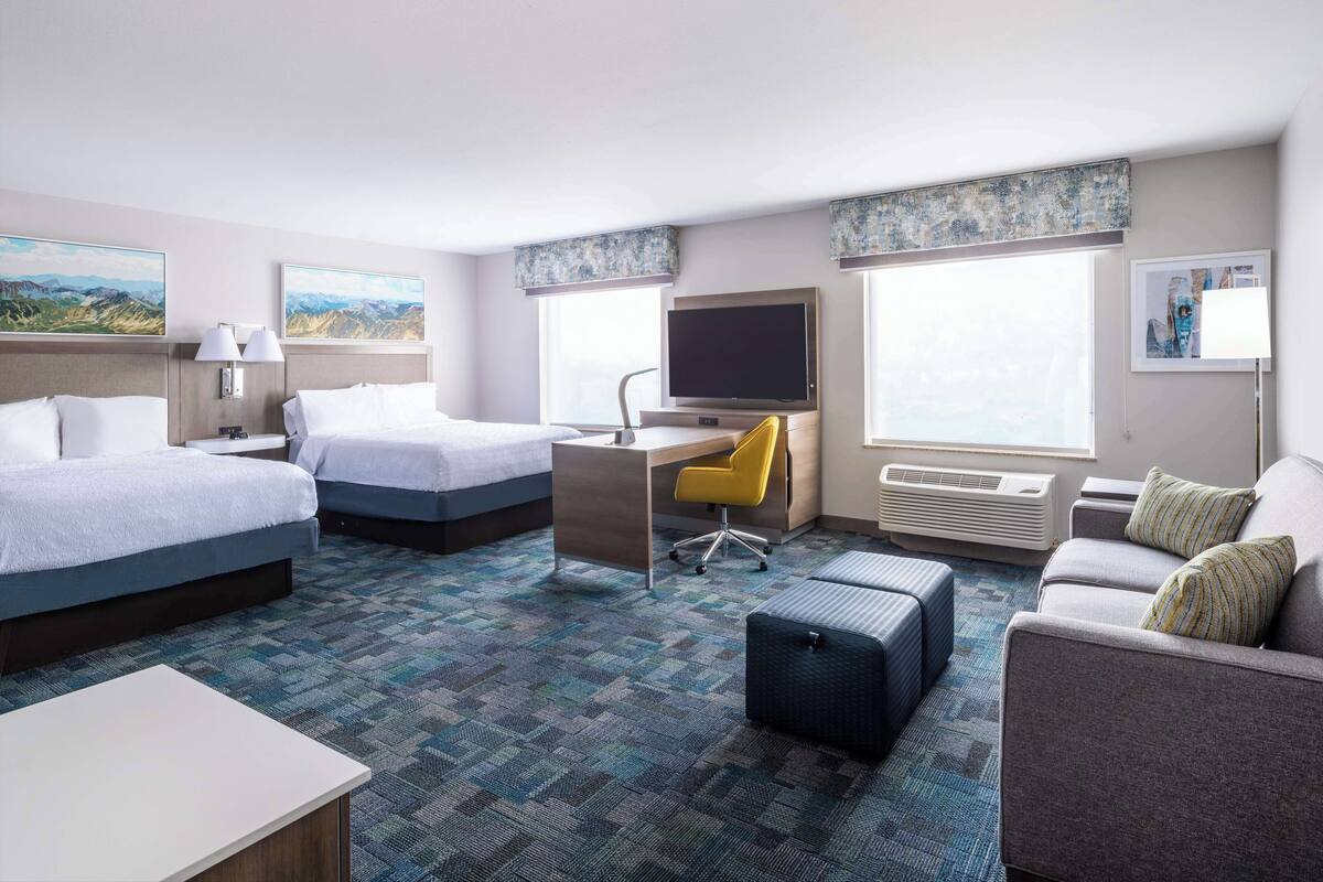 A hotel room with two beds, a desk with a computer, a chair, a sofa, and a large window.