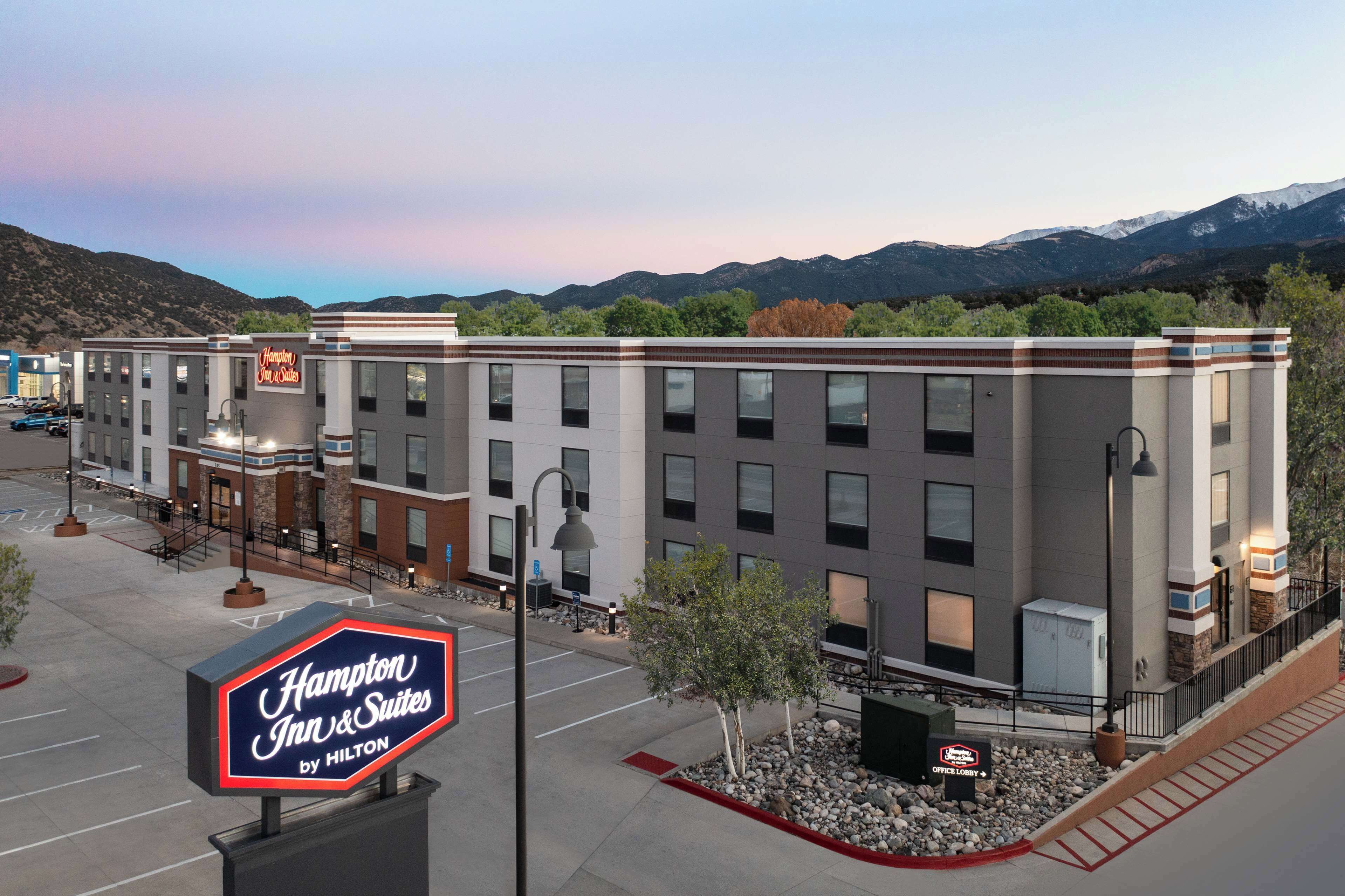 A Hampton Inn & Suites by Hilton hotel with a parking lot, a signboard, and a view of mountains in the background.