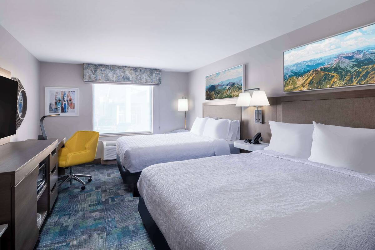 A hotel room with two beds, a desk, a chair, and a large window.