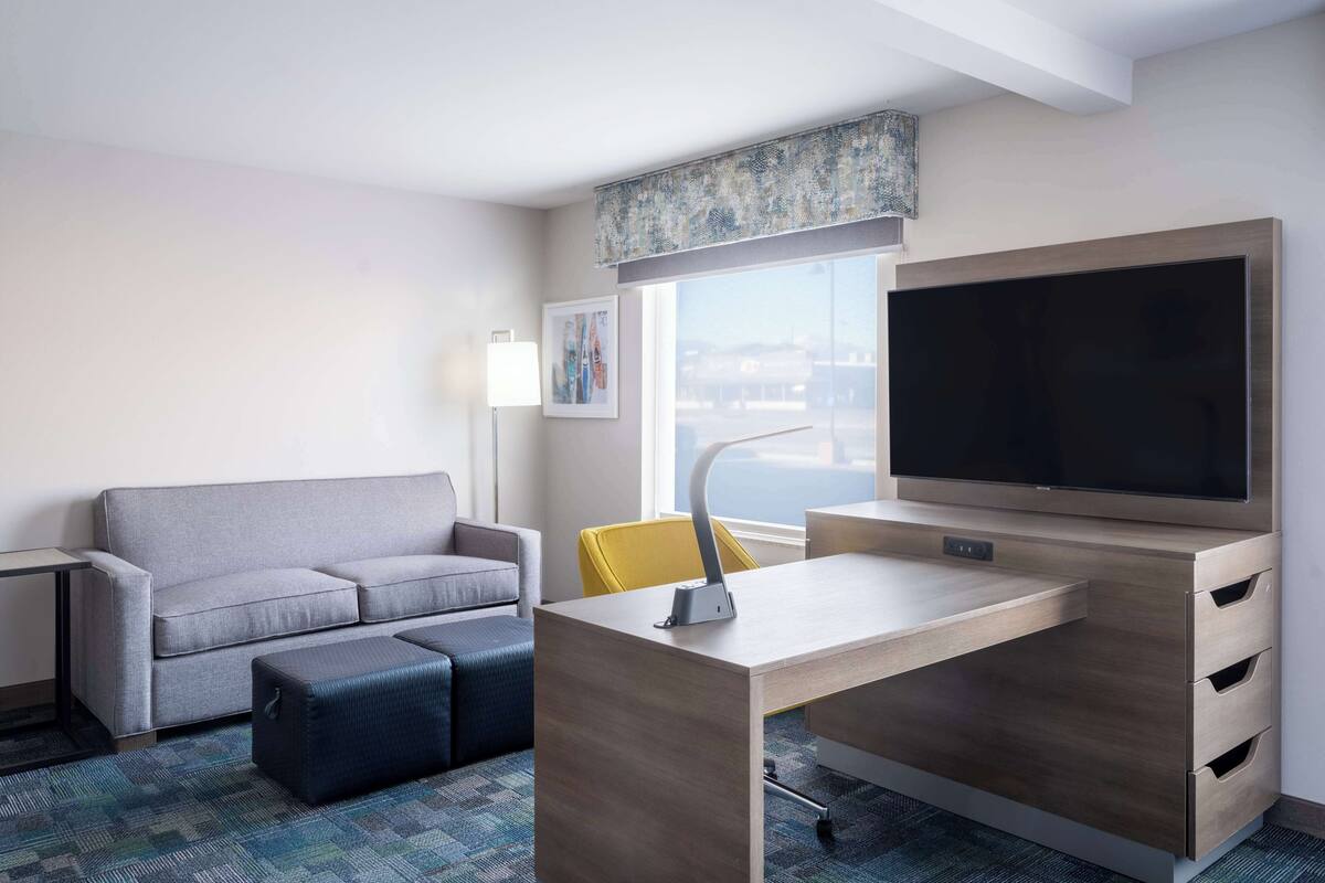 A hotel room with a grey sofa, a wooden desk with drawers, a flat-screen TV, a yellow chair, a lamp, and a window with a view.