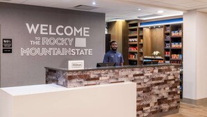 A reception desk with a "Welcome to the Rocky Mountain State" sign, a Hilton Honors logo, and a variety of snacks on shelves behind the counter.