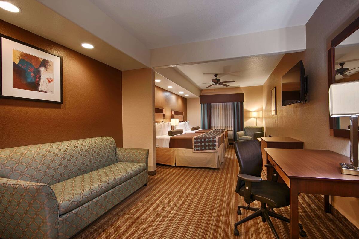 Ein Hotelzimmer mit einer gepolsterten Couch, einem Schreibtisch mit Lampe, einem Sessel, einem Bett mit gemustertem Überwurf, einem Deckenventilator und einem gerahmten Bild an der Wand.