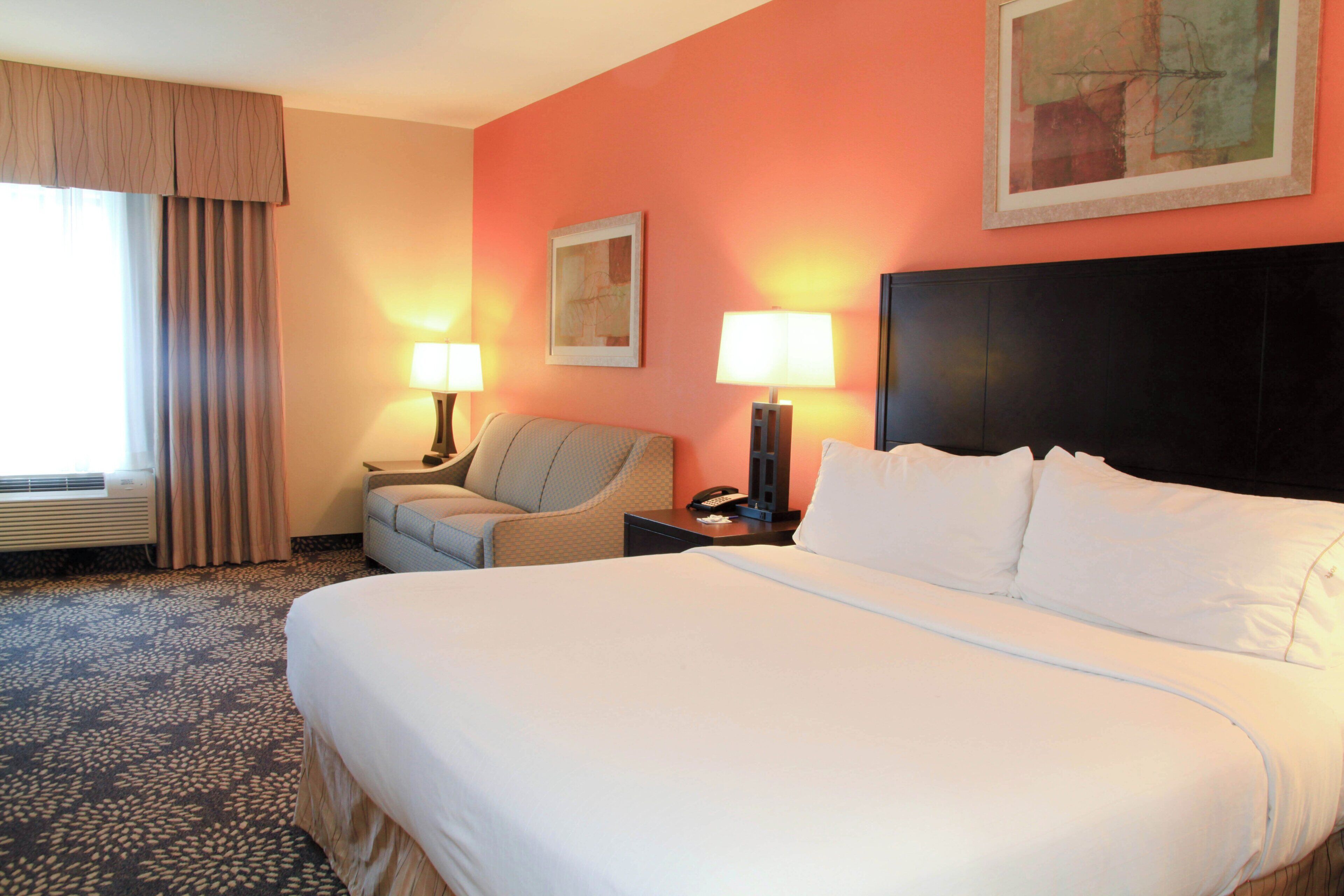 Una habitación de hotel con una cama grande, dos lámparas de noche, una silla, un escritorio con teléfono y una ventana con cortinas.