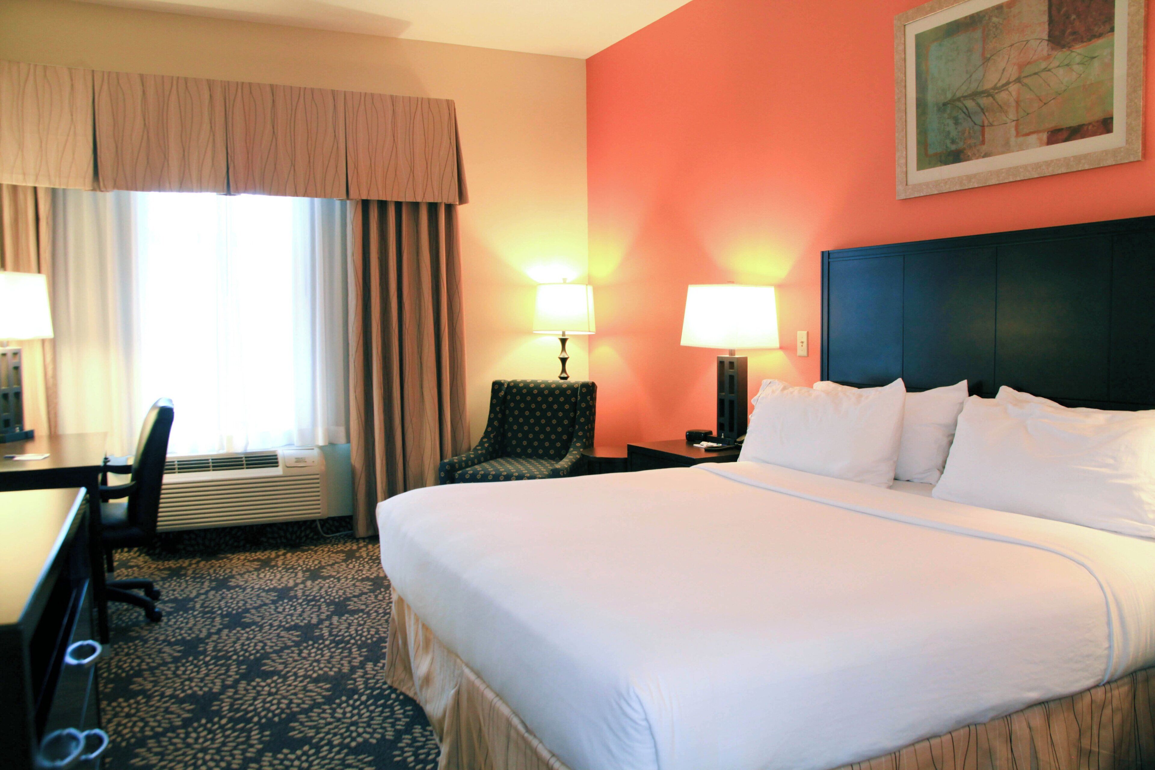 A hotel room with a large bed, two bedside lamps, a desk with a chair, and a window with curtains.