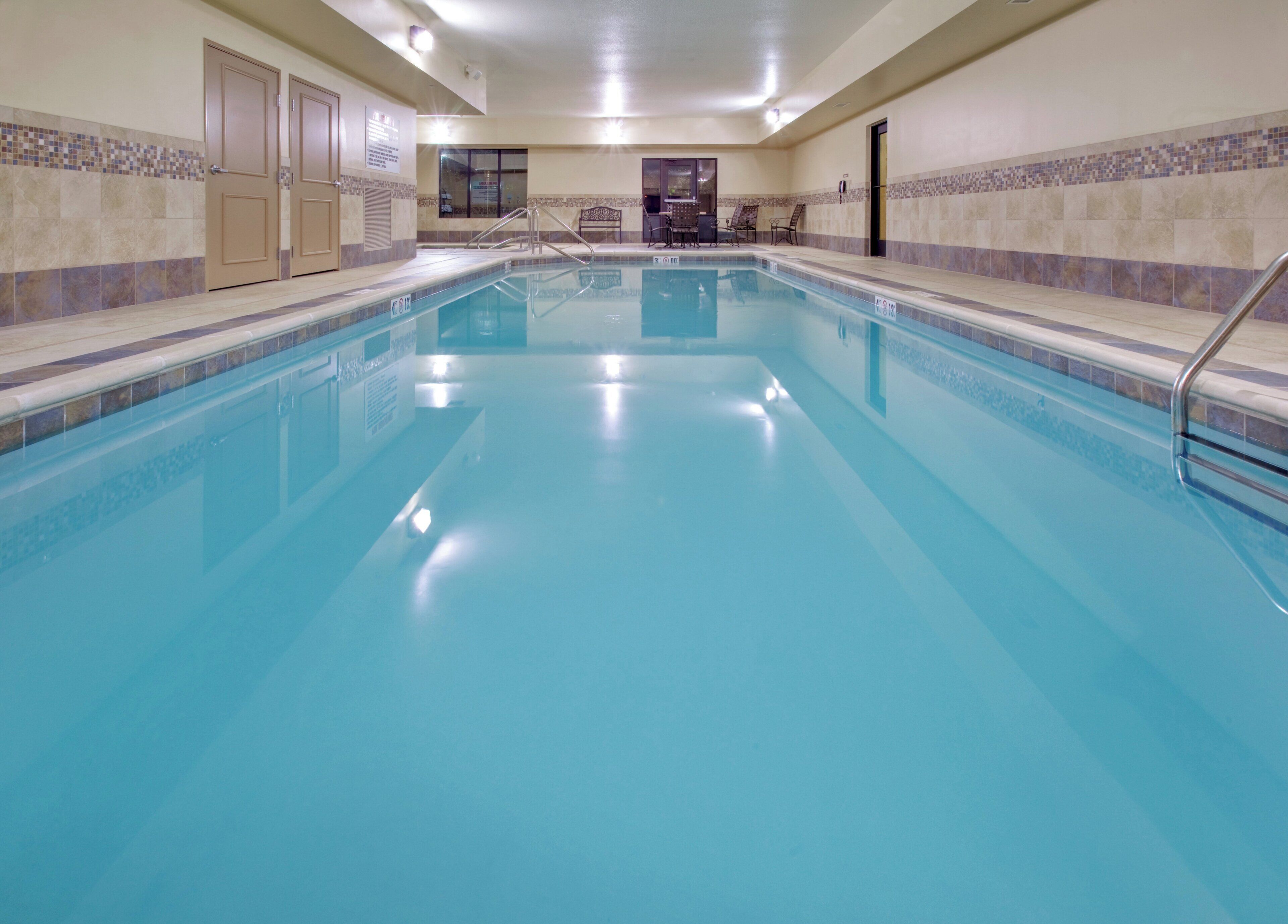 A large indoor swimming pool with clear blue water, surrounded by tiled walls and a bench area with chairs.