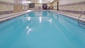 A large indoor swimming pool with clear blue water, surrounded by tiled walls and a bench area with chairs.