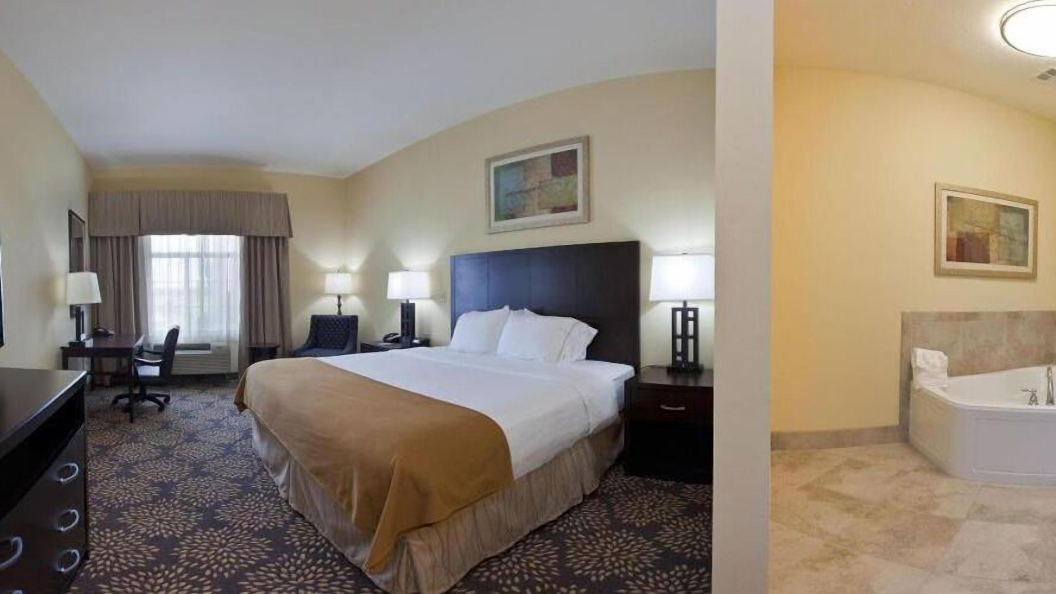 Une chambre d’hôtel avec un grand lit, un bureau, une chaise, une lampe de bureau, une table de chevet, un tableau au mur et une salle de bain avec une baignoire.
