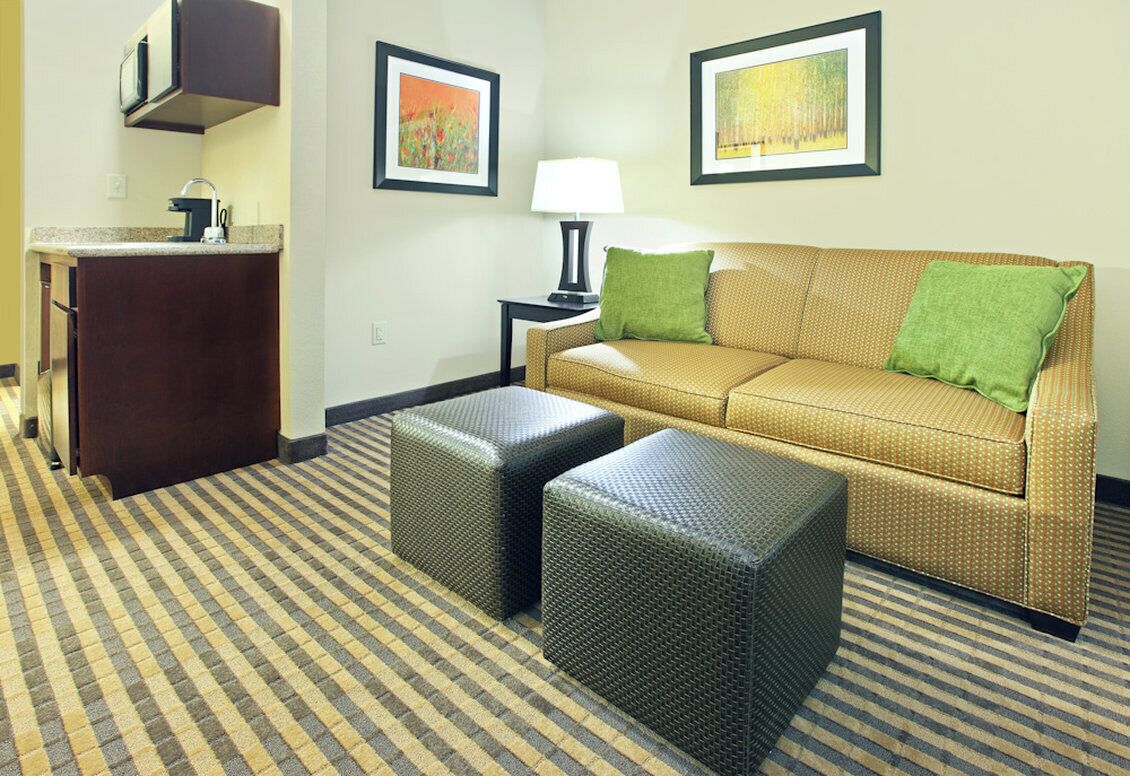 Ein Hotelzimmer mit einer Couch, zwei Hocker, einem kleinen Tisch mit Lampe, einer Küchenzeile und Bildern an den Wänden.