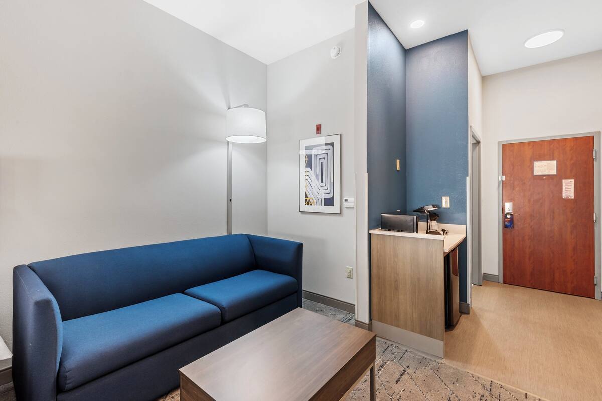 Un hall d’hôtel moderne avec un canapé bleu, une table en bois, un bureau avec un ordinateur, une œuvre d’art encadrée et une porte en bois.