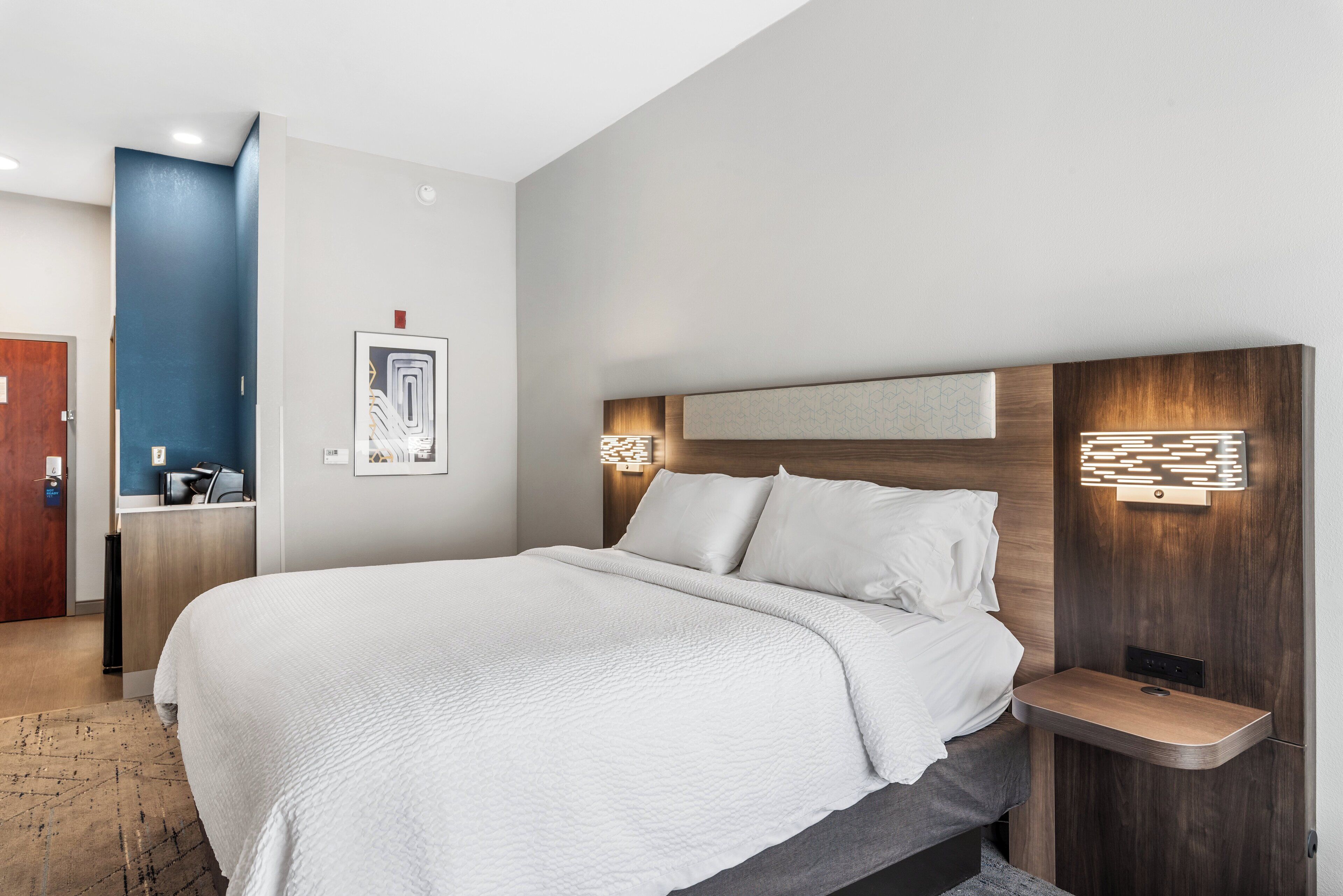 Quarto de hotel moderno com uma cama grande, uma mesa de cabeceira e luminária fixada na parede.