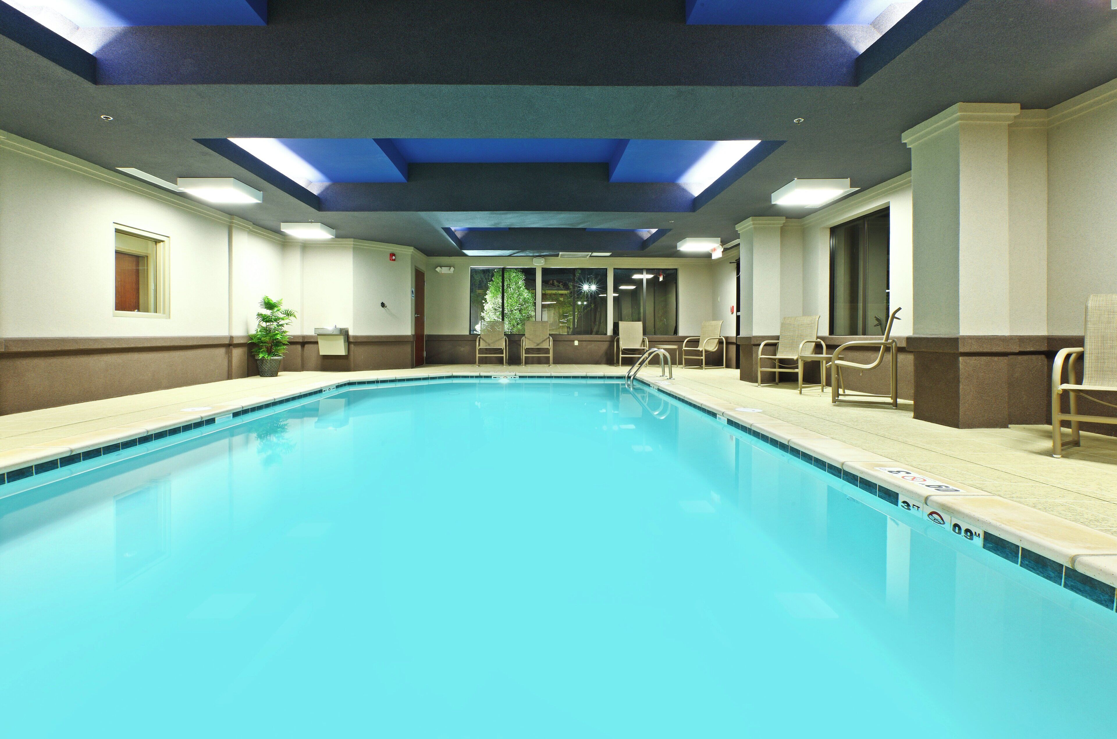 Una gran piscina interior con superficie de agua azul cristalina, rodeada por una zona de piscina con sillas y una planta en maceta.