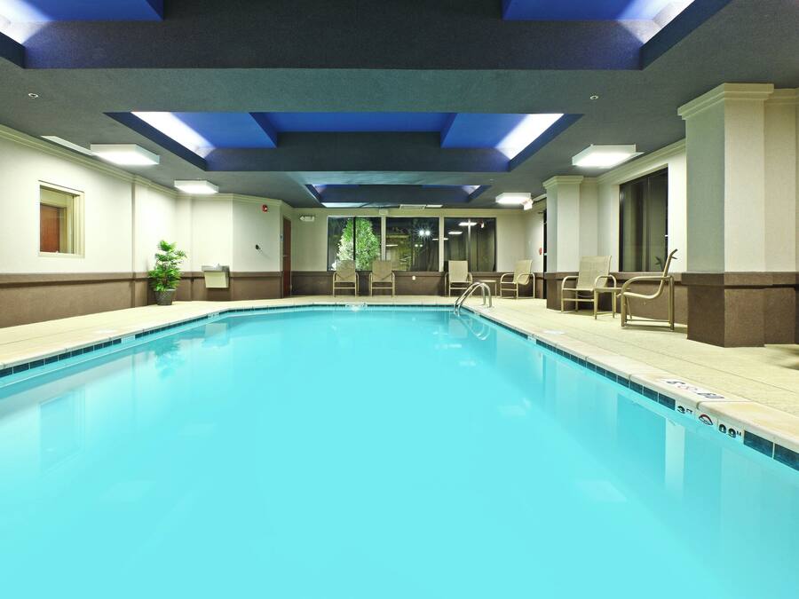 Una gran piscina interior con superficie de agua azul cristalina, rodeada por una zona de piscina con sillas y una planta en maceta.