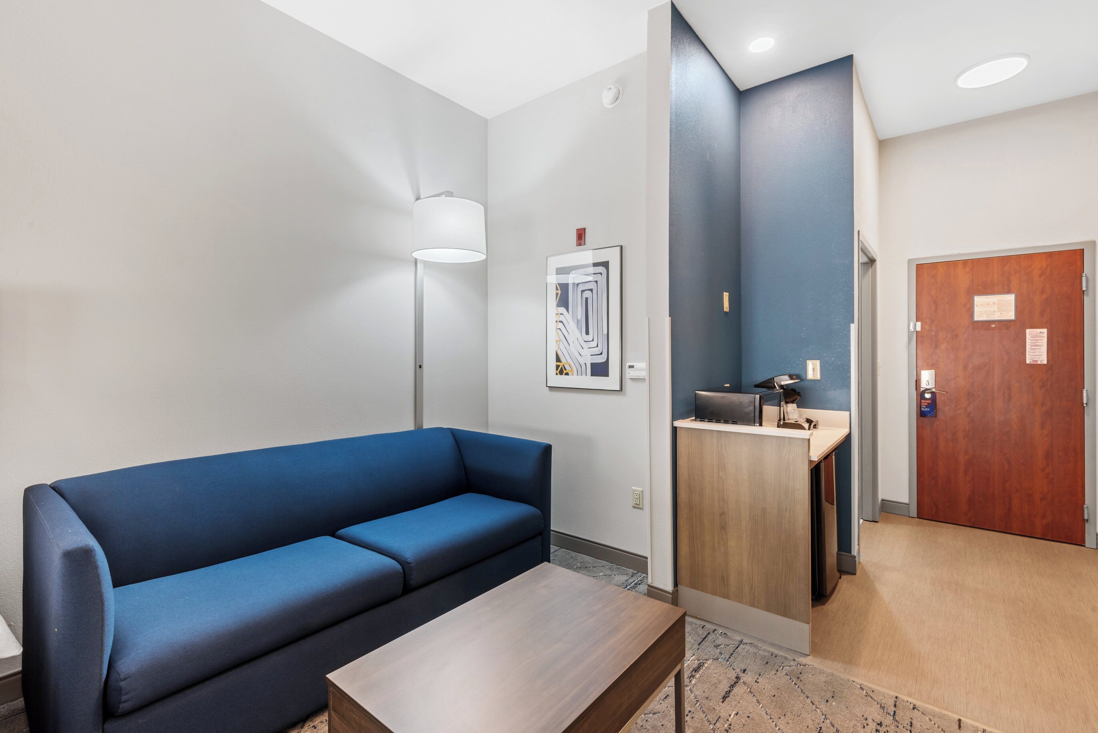 A modern hotel lobby with a blue sofa, a wooden table, a desk with a computer, a framed artwork, and a wooden door.