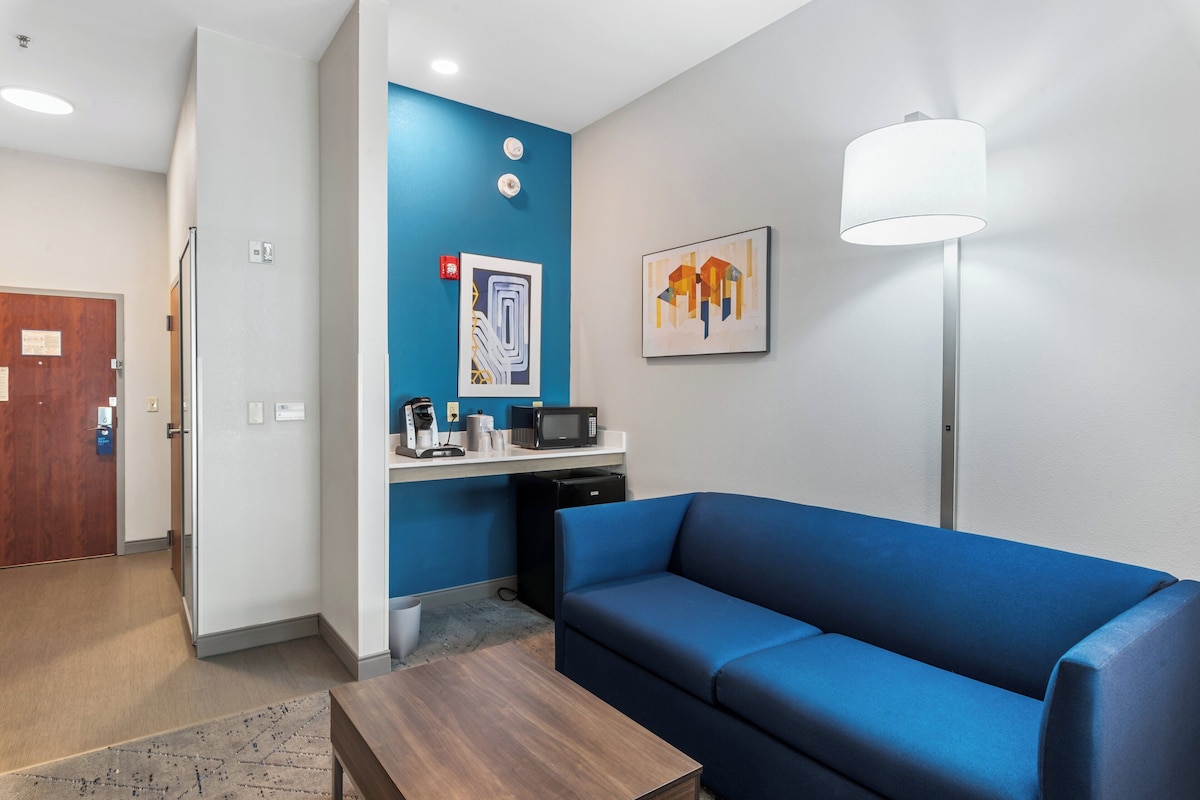 A modern hotel room with a blue sofa, a wooden coffee table, a microwave, and a small kitchenette.