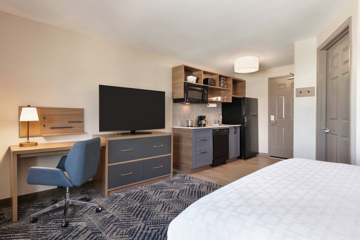 Ein Hotelzimmer mit einem Bett, einem Schreibtisch mit Stuhl, einem Fernseher und einer Küchenzeile inklusive Mikrowelle und Kühlschrank, sowie einer Tür.