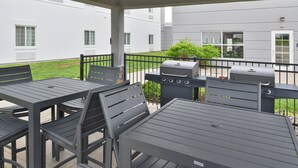 Een terras buiten met metalen tafels en stoelen, een barbecue en een overdekte doorgang.