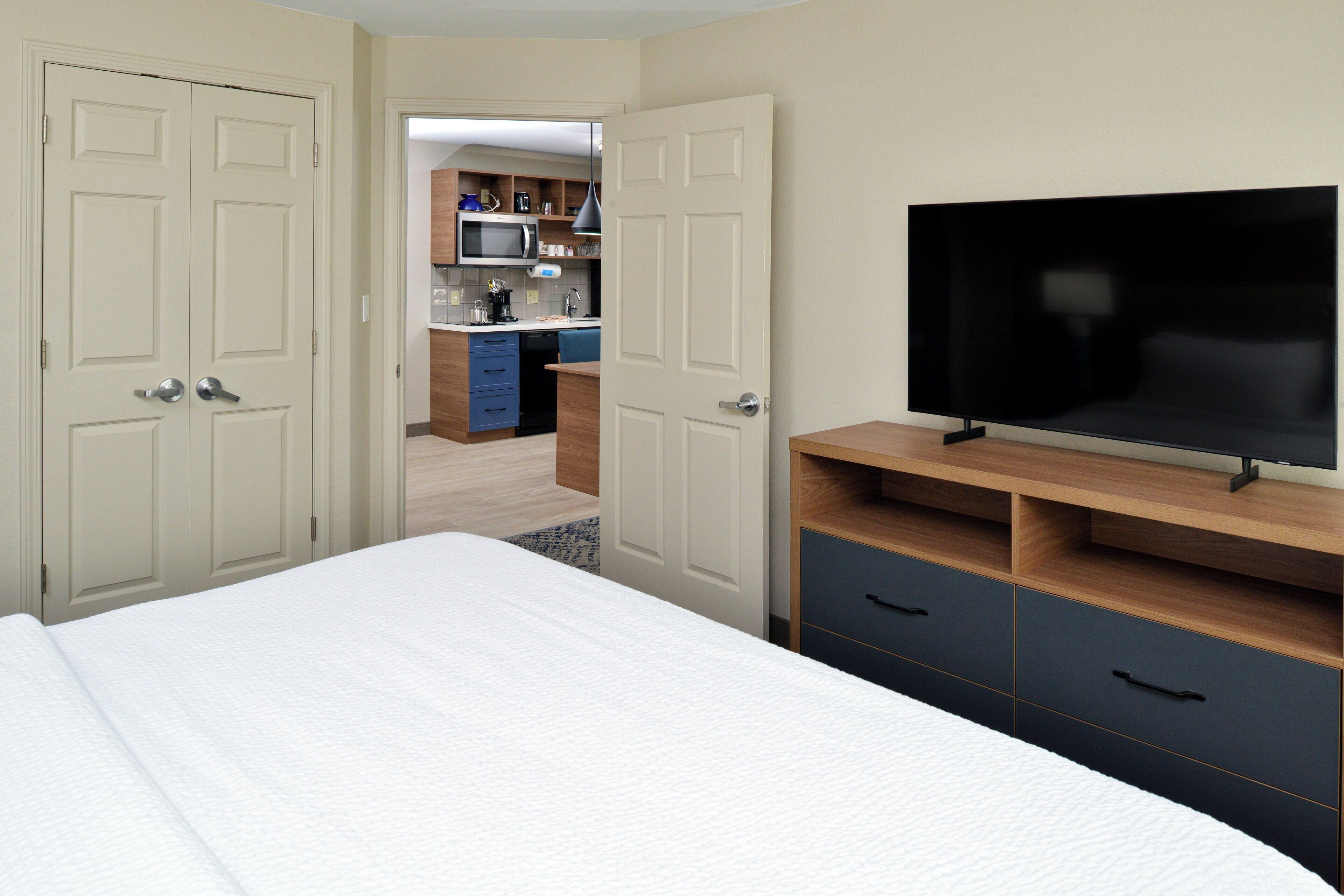 A hotel room with a bed, a TV, and a kitchenette in the background.