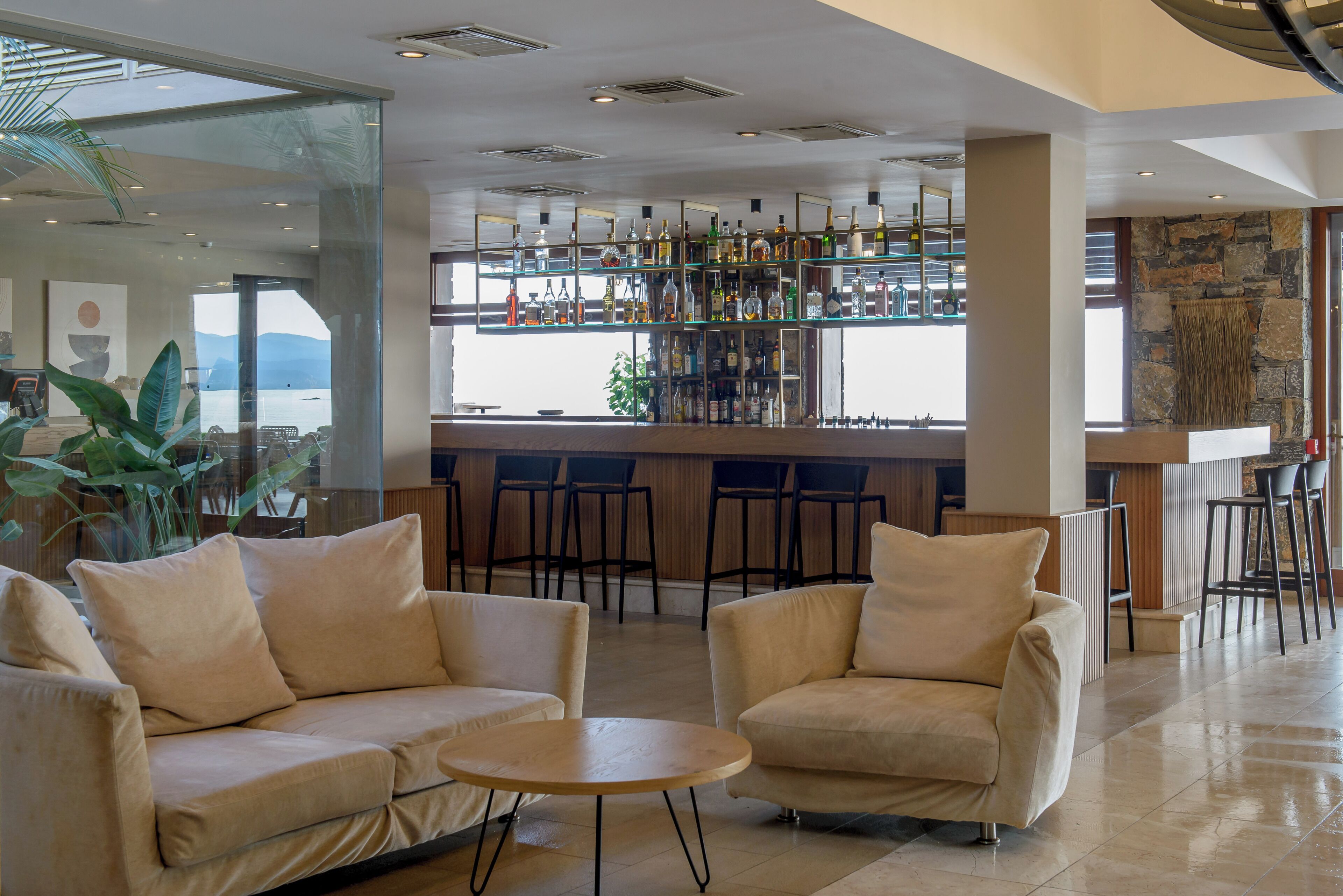 A bar area with a variety of bottles, a seating area with two sofas and a small table, and a view through large windows.