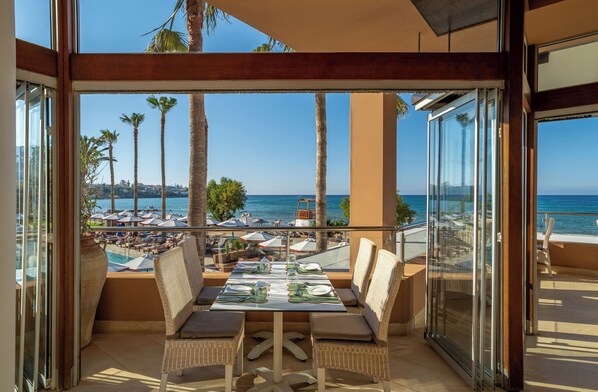 A balcony with a table set for dining, overlooking a beach with palm trees and umbrellas.