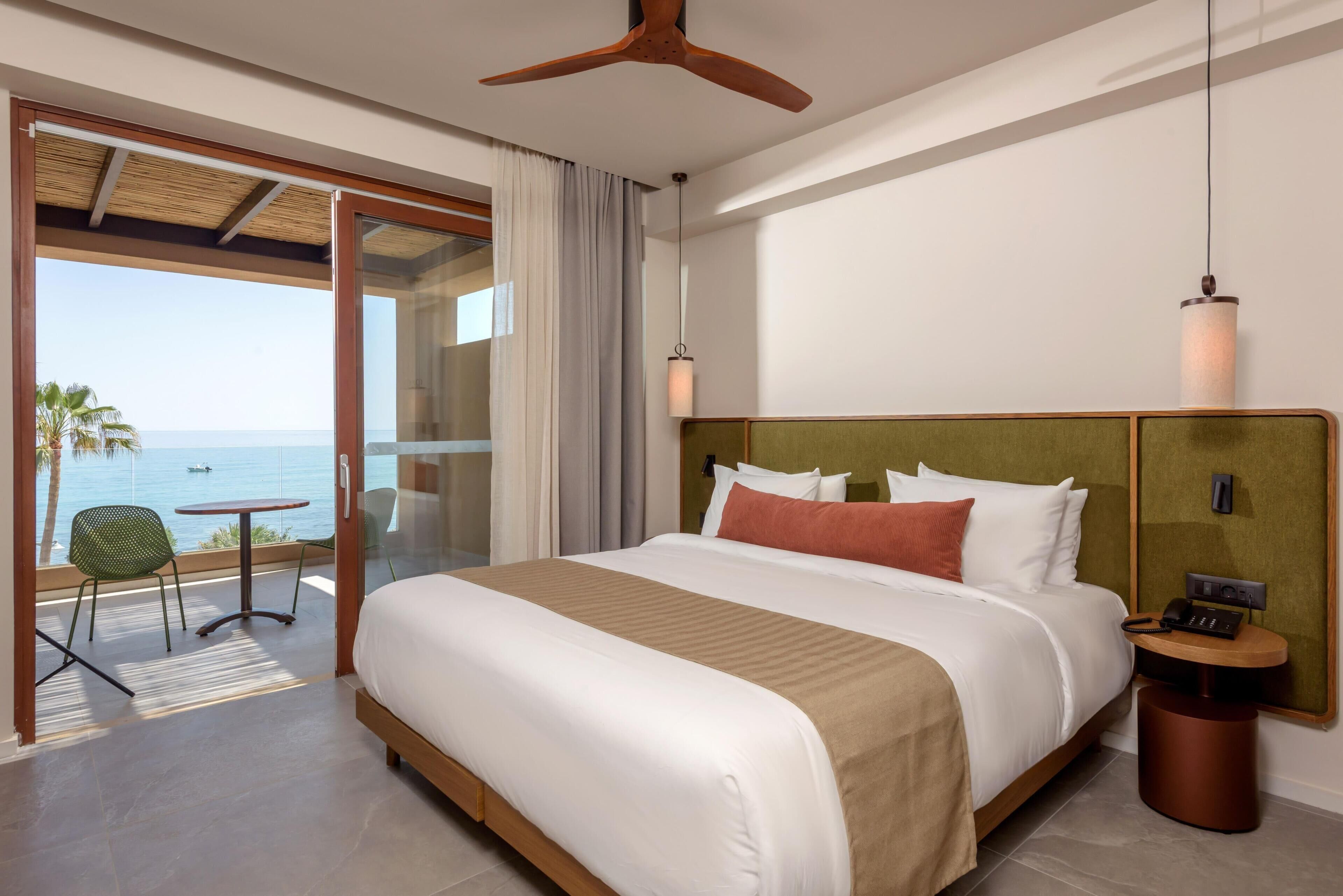 Habitación de hotel con cama, una mesa, una silla, ventilador de techo y vista al océano.