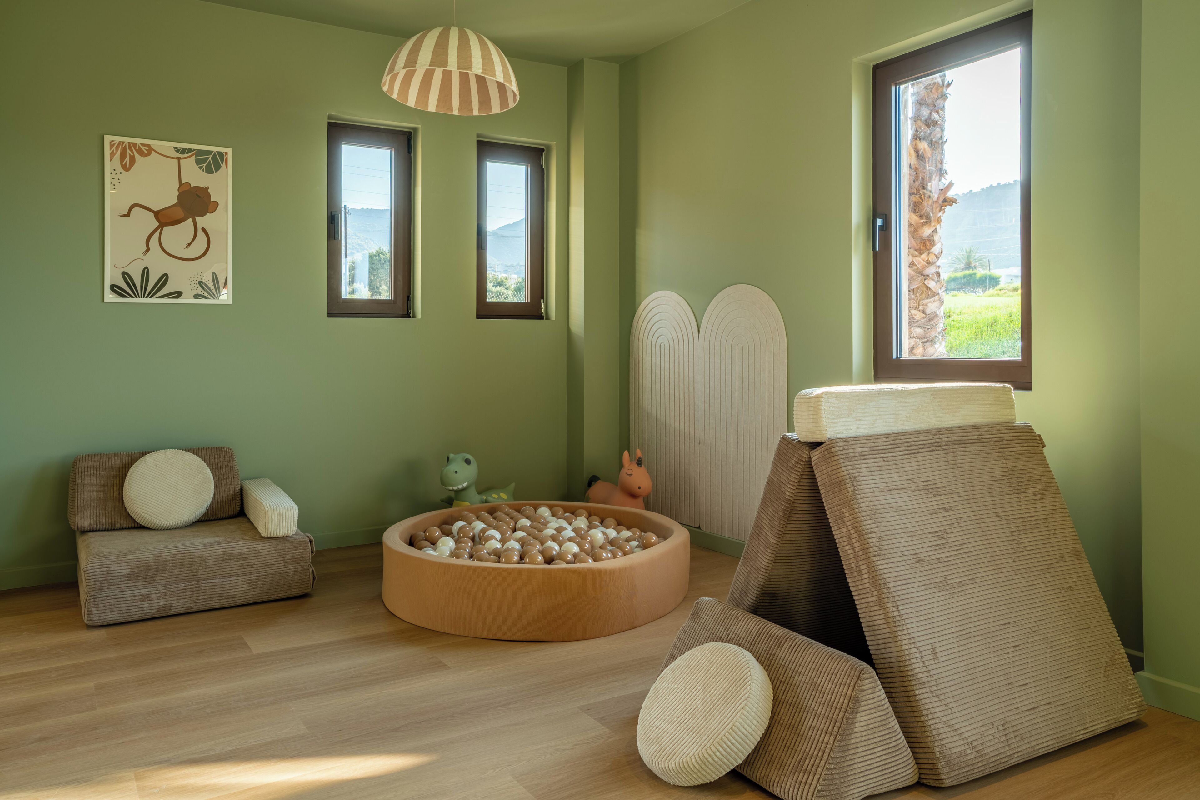 A room with a wooden floor, a round play area with balls, a sofa with cushions, a hanging lamp, and a window with a view.