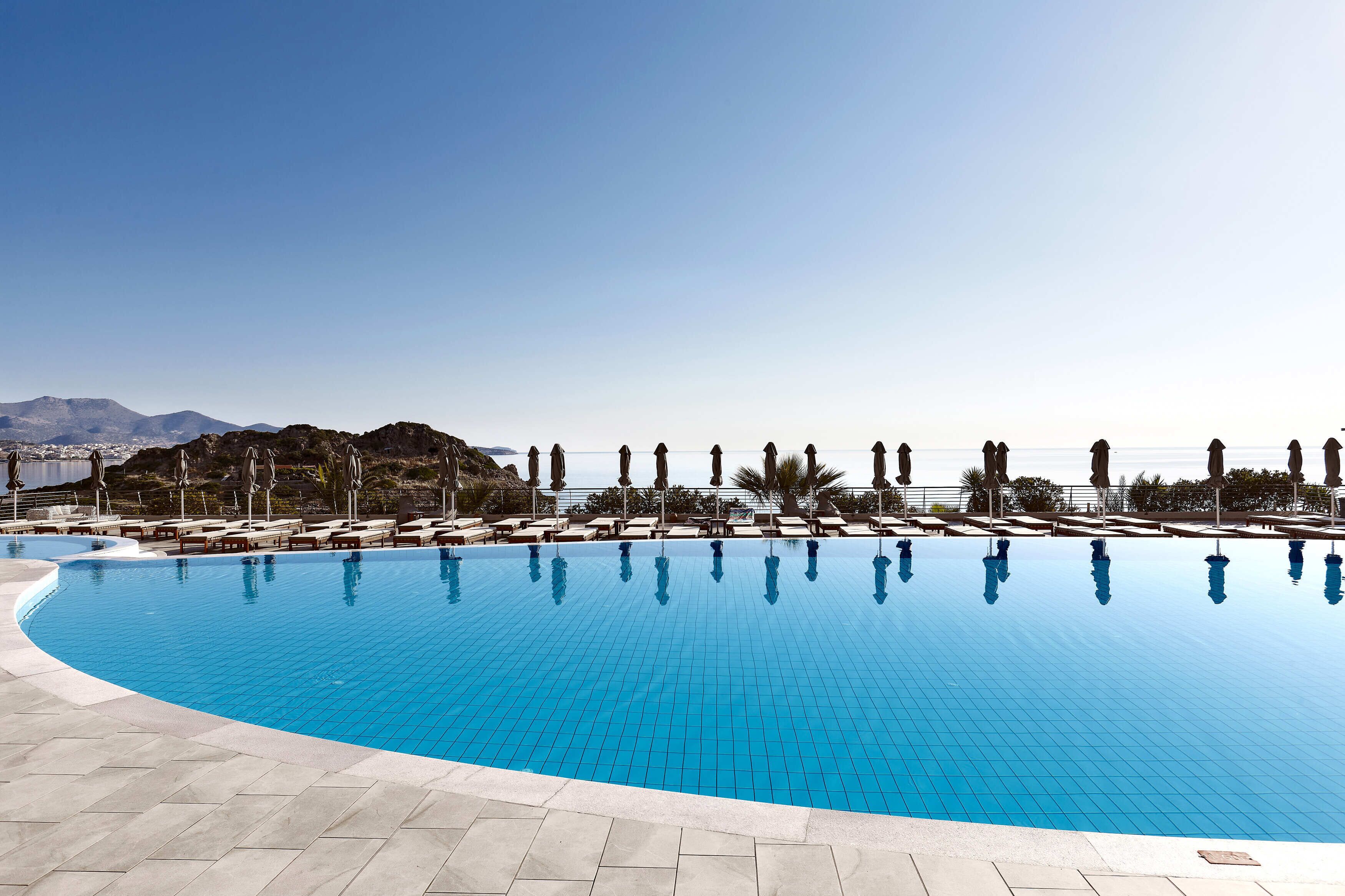 Une grande piscine d’un bleu éclatant, bordée de chaises longues et d’ombrelles, et entourée de montagnes sous un ciel dégagé.