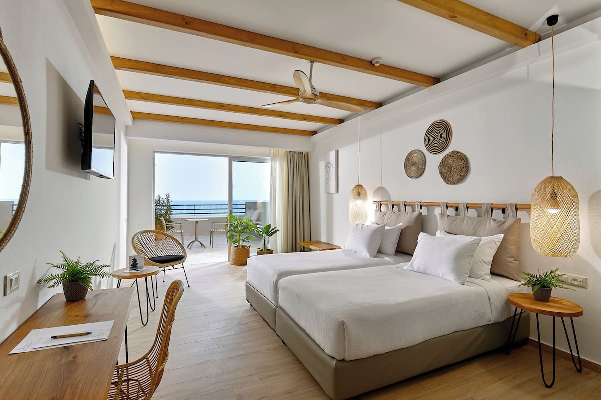A modern bedroom with a large bed, bedside tables, a desk, and a chair. There are potted plants and a view of the outdoors.