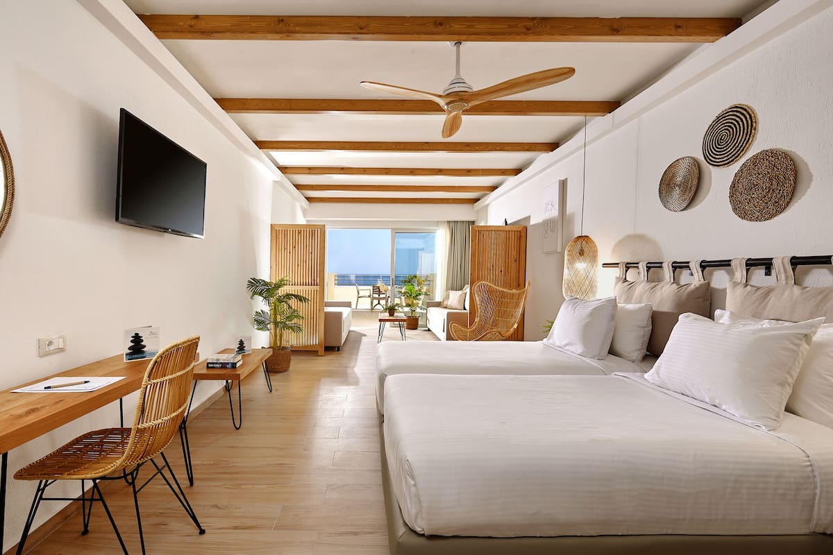 A modern hotel room with a wooden ceiling, a flat-screen TV, a bed with white linens, a desk with a chair, and a view of the sea.