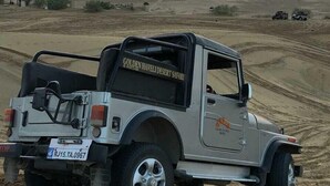 Een zilveren terreinwagen met de tekst 'Golden Haveli Desert Safari' op de zijkant, geparkeerd op een zanderig terrein met een ondergaande zon op de achtergrond.