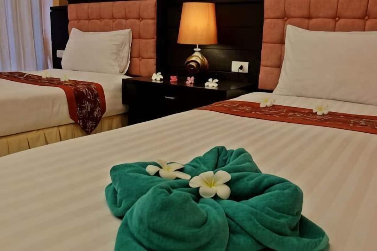 Quarto de hotel com duas camas, mesa de cabeceira com abajur e arranjo floral sobre a cama.