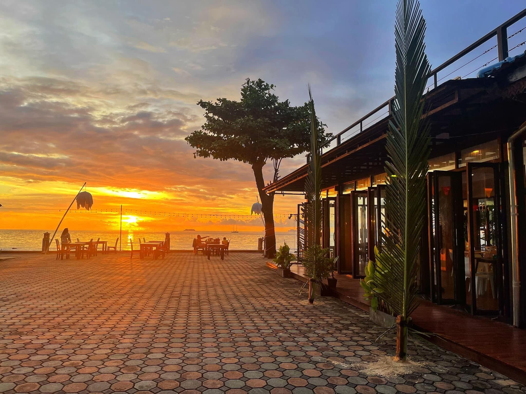 A sunset view from a terrace with hexagonal tiles, tables, chairs, and a palm tree.