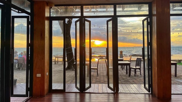 A room with large glass doors leading to an outdoor seating area with tables and chairs, overlooking a beach at sunset. - Andaman Lanta Resort (Ko Lanta)