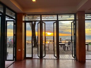 Une pièce dotée de grandes portes vitrées donnant sur un espace extérieur aménagé avec des tables et des chaises, et offrant une vue sur une plage au coucher du soleil.