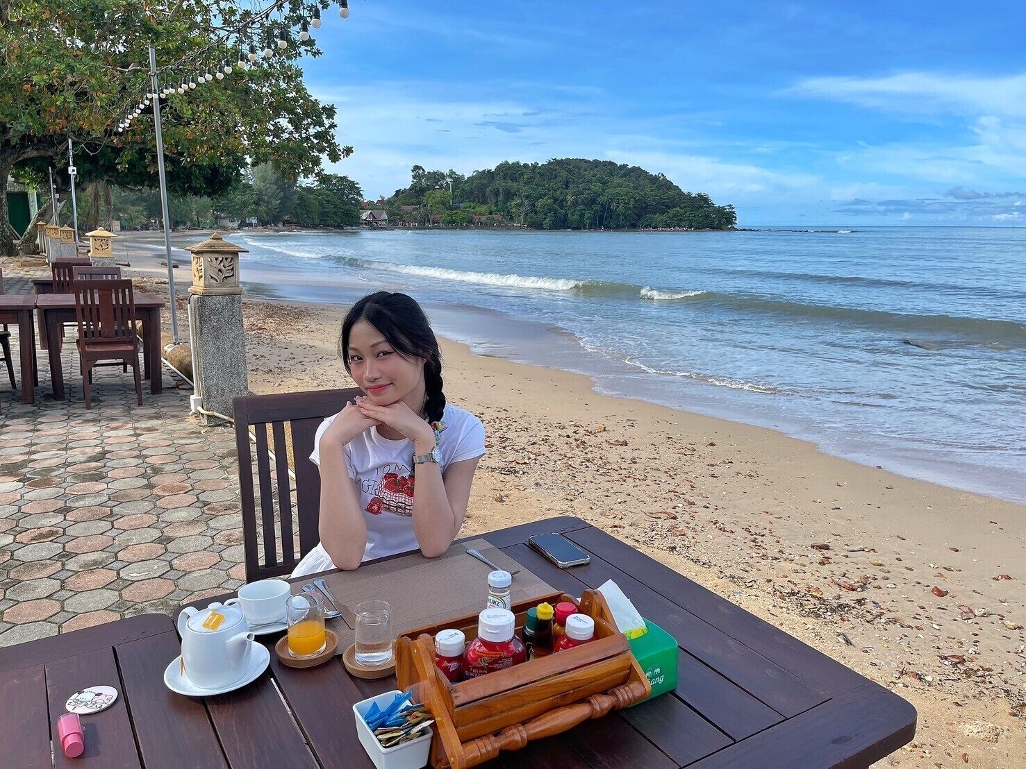 Una mesa junto a la playa con comida dispuesta, incluyendo un calentador de té, tazas y condimentos, con vista al océano.