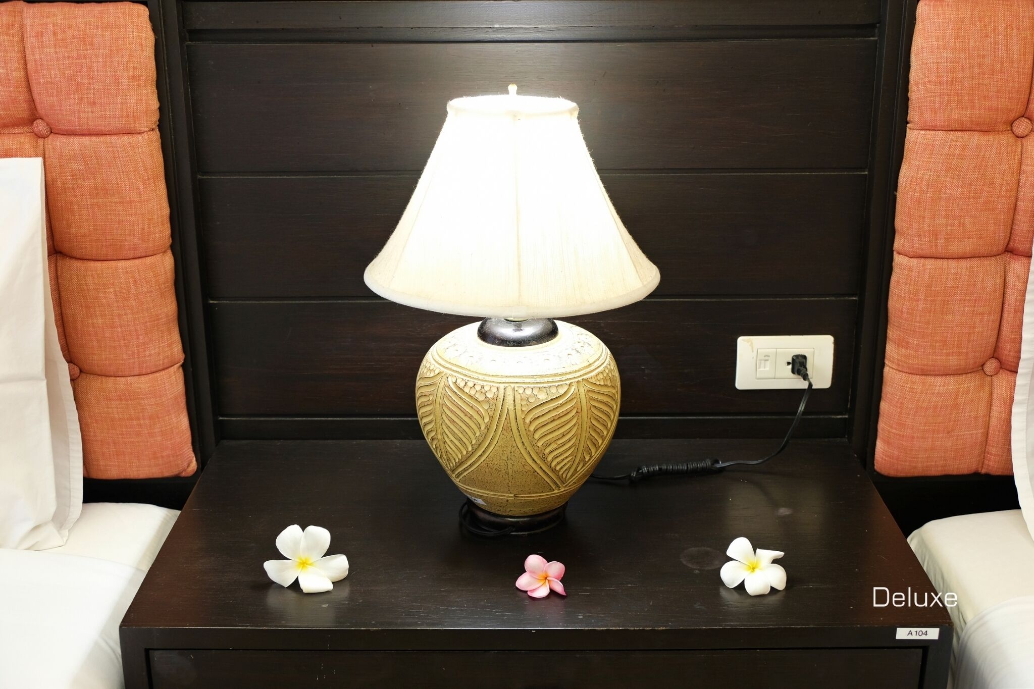 A bedside table with a decorative lamp, two pillows, and a flower.